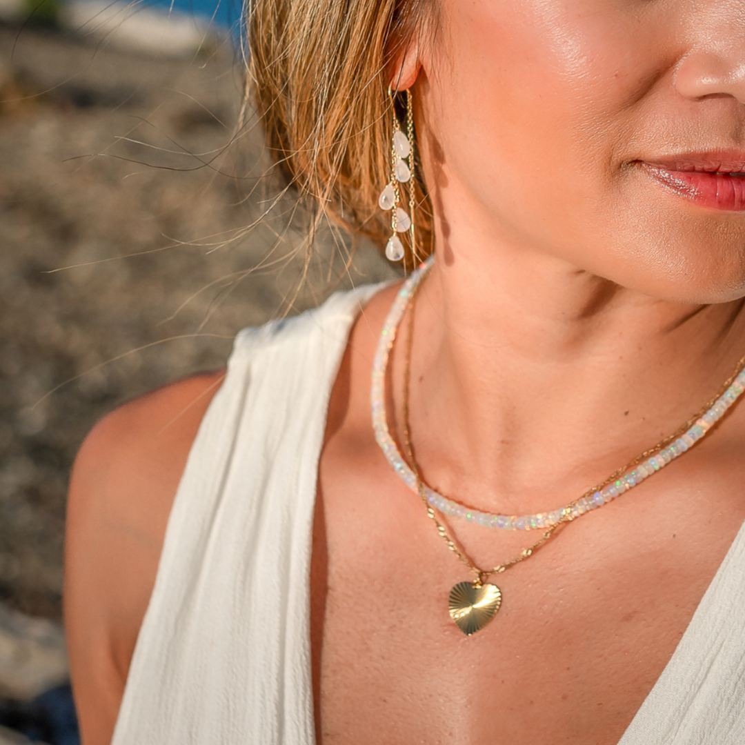 Woman wearing a gold heart-shaped necklace and earrings with a blurred beach background