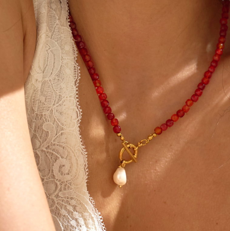 Necklace with red beads and a gold toggle clasp and pearl pendant worn by a person in white lace garment.