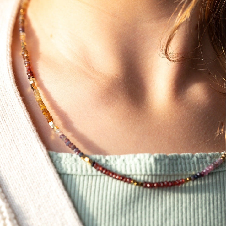 A close-up lifestyle shot of a person wearing a delicate sapphire beaded necklace with faceted gemstone beads in amber, red, purple, and gold tones, fall necklace September birthstone necklace 