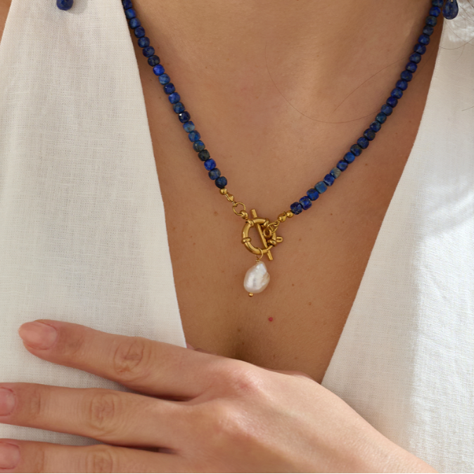 Person wearing a blue beaded gemstone necklace with a gold pendant and pearl accent, against a white background.