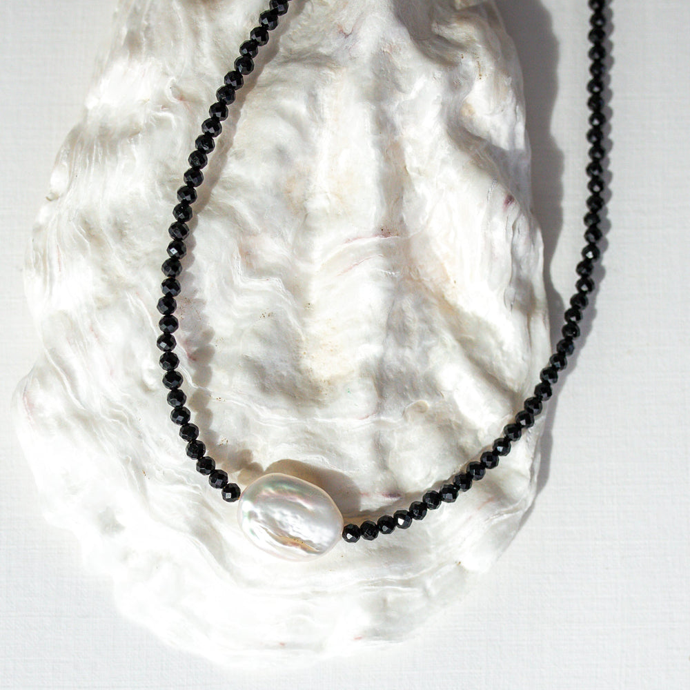 A beaded necklace with small black faceted beads and a large, irregular white pearl-like bead at the center, displayed on a white textured shell against a white background.