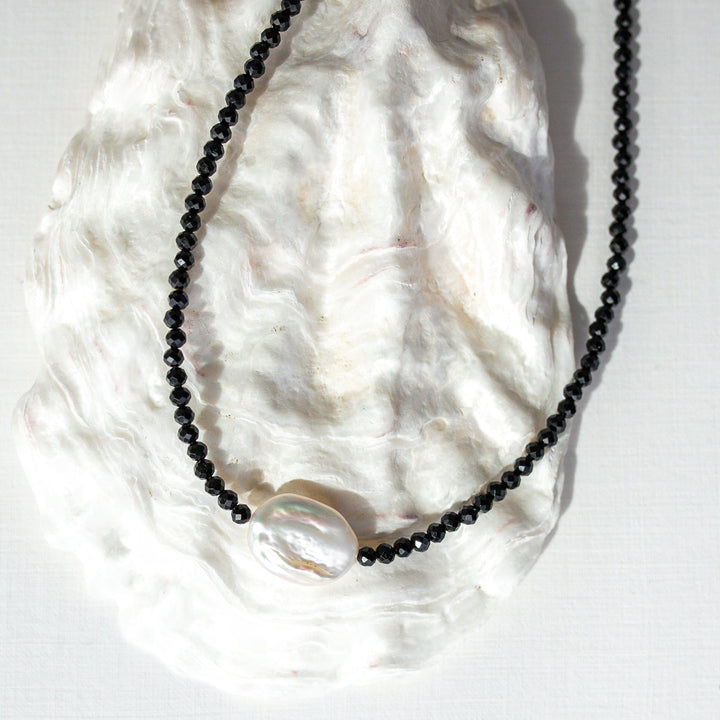 A beaded necklace with small black faceted beads and a large, irregular white pearl-like bead at the center, displayed on a white textured shell against a white background.
