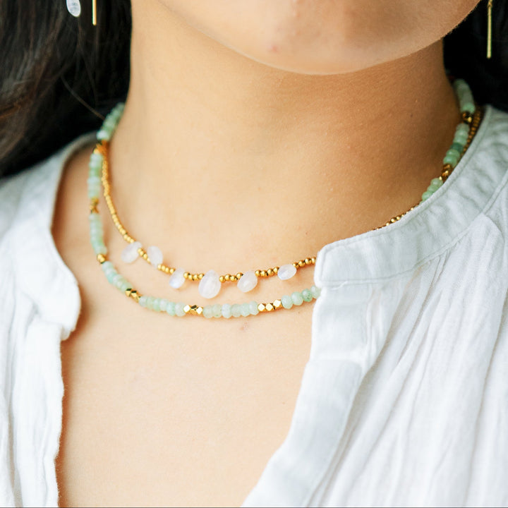 Close-up of a person wearing a gold moonstone necklace layered with  green adventurine beaded necklace with earrings.