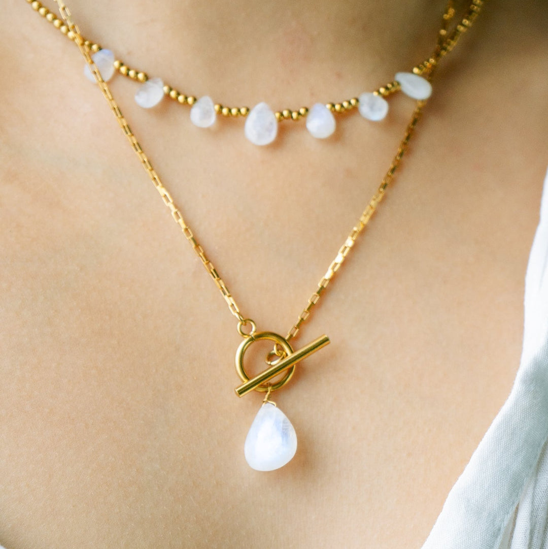 Gold necklace with a toggle clasp and teardrop-shaped pendant on a person wearing a white top.