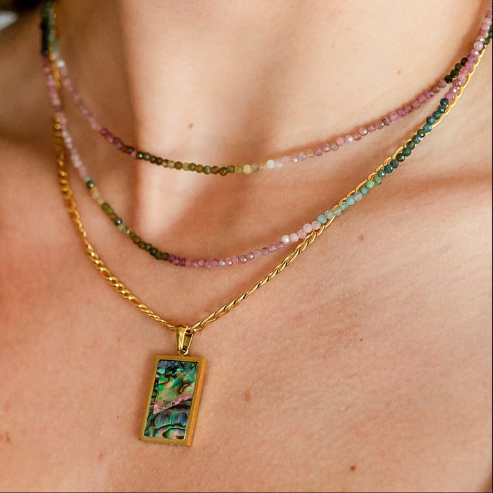 Close-up of a person wearing multiple tourmaline necklaces with a focus on one gold necklace with a green shell pendant. Waterproof Gold Figaro Chain 