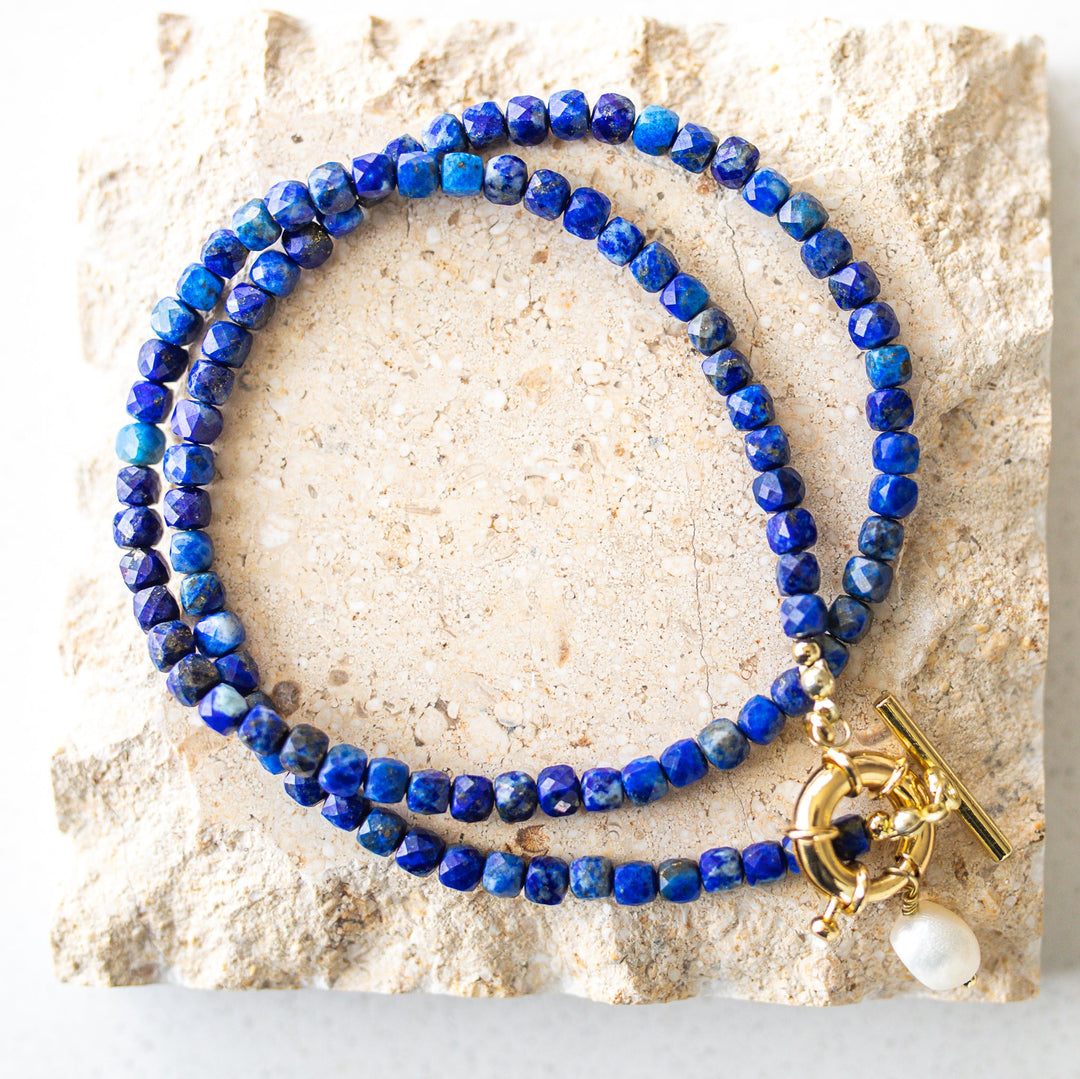 Blue beaded necklace with a gold clasp and pearl charm on a stone surface