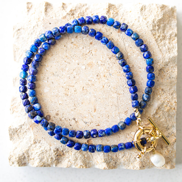 Blue beaded necklace with a gold clasp and pearl charm on a stone surface